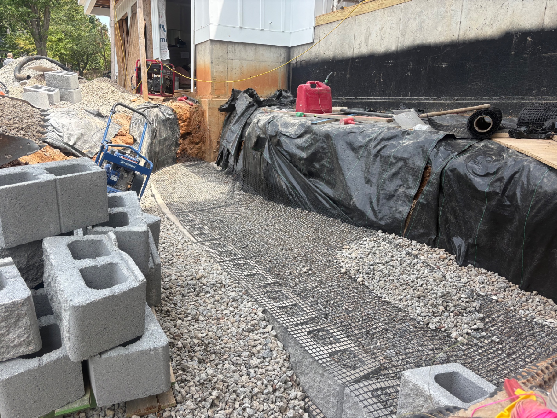 Compacted stone retaining wall base with geogrid reinforcement and filter fabric beside a home foundation