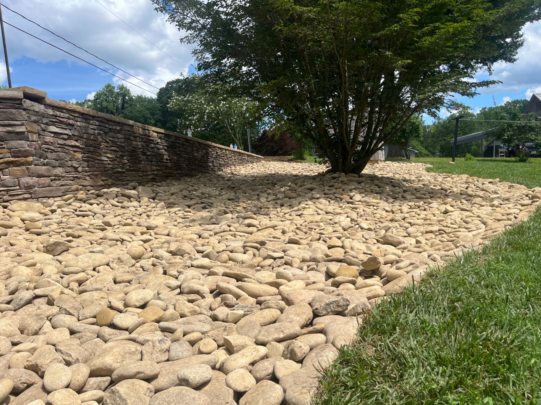 Large river-rock drainage bed on a slope beside a stone retaining wall with a mature tree in Terrell, NC