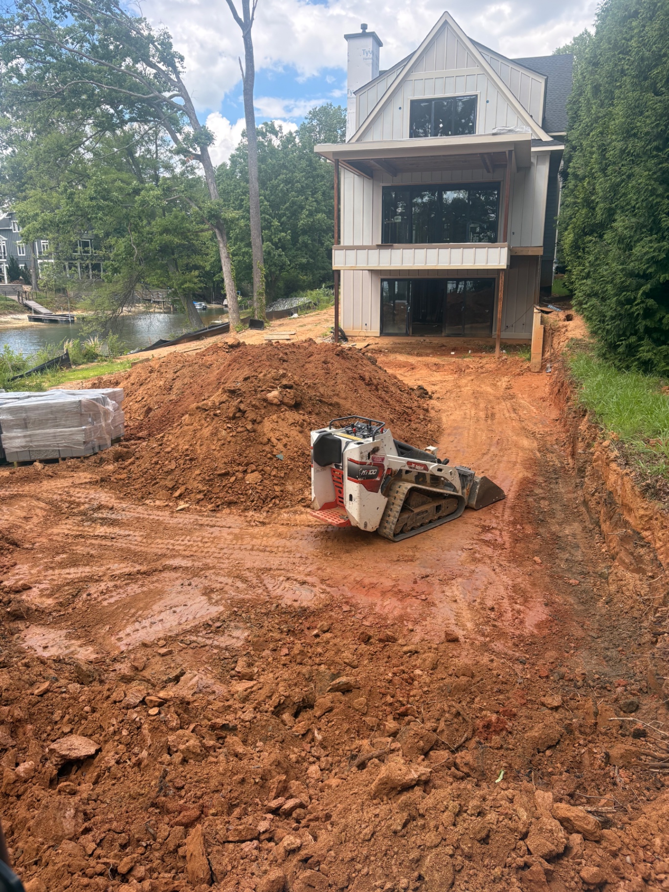 Mini track loader moving excavated clay behind a new lakefront home under construction in Cornelius, NC, with pavers staged for the next phase